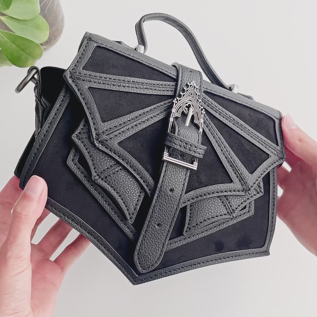Bolso de mano pequeño estilo gótico con inspiración en los murciélagos sujetado por unas manos que enseñan sus detalles en el tejido y las hebillas metálicas