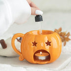 Quemador de aceites esenciales y calentador de cera con forma de taza de calabaza con nata montada encima estilo halloween otoñal vista posterior poniendo aceite en la parte superior y vista de la vela de te encendida sobre escritorio con decoración al rededor.