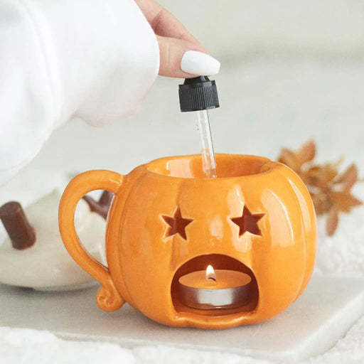 Quemador de aceites esenciales y calentador de cera con forma de taza de calabaza con nata montada encima estilo halloween otoñal vista posterior poniendo aceite en la parte superior y vista de la vela de te encendida sobre escritorio con decoración al rededor.