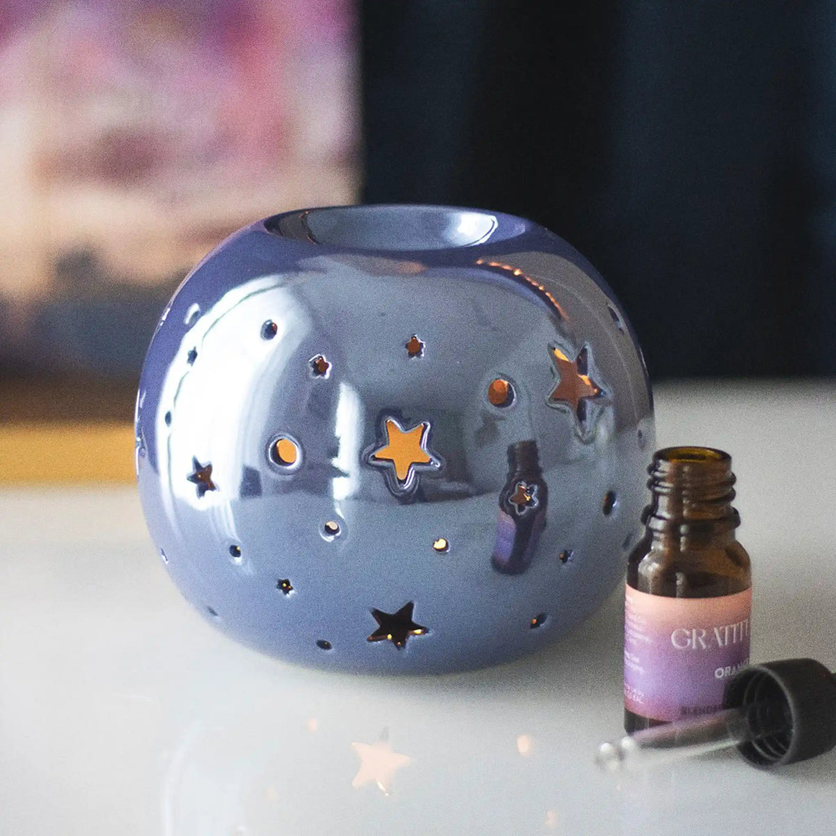 Quemador de aceite en forma de bola con estrellas perforadas en color morado iridiscente vista frontal sobre escritorio con decoración al rededor.