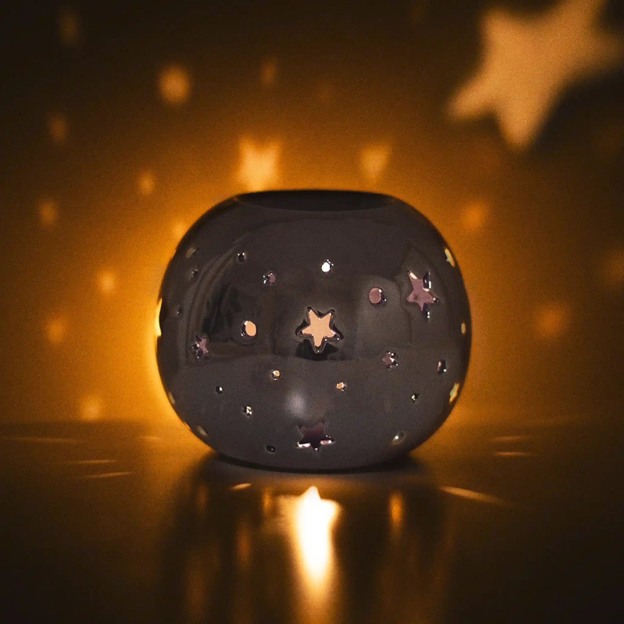Quemador de aceite en forma de bola con estrellas perforadas en color morado iridiscente vista sobre escritorio con luz apagada y vela encendida.
