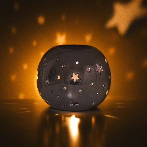 Quemador de aceite en forma de bola con estrellas perforadas en color morado iridiscente vista sobre escritorio con luz apagada y vela encendida.
