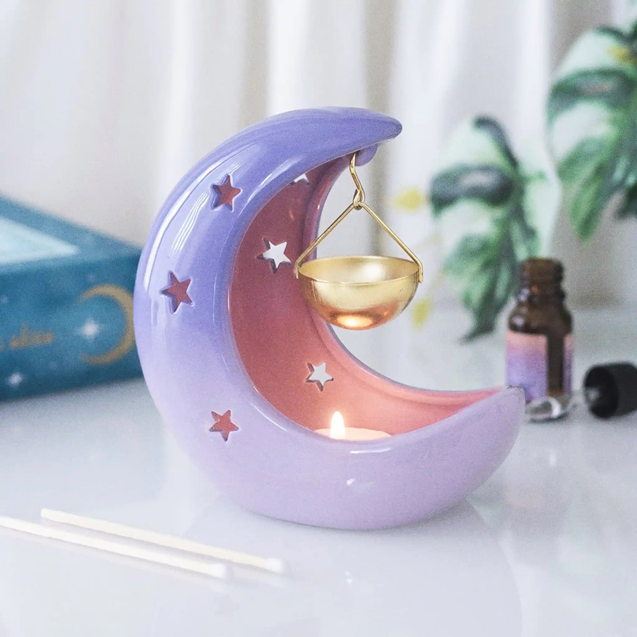 Quemador de aceites esenciales en forma de luna creciente con colores violetas degradados y recipiente colgante para aceite en color dorado vista frontal sobre escritorio con decoración al rededor.