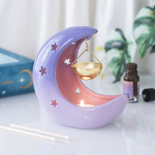 Quemador de aceites esenciales en forma de luna creciente con colores violetas degradados y recipiente colgante para aceite en color dorado vista frontal sobre escritorio con decoración al rededor.