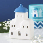Quemador de aceites esenciales y calentador de cera con forma de casa de Santorini inspirado en Grecia en blanco y azul vista frontal sobre escritorio con decoración al rededor.