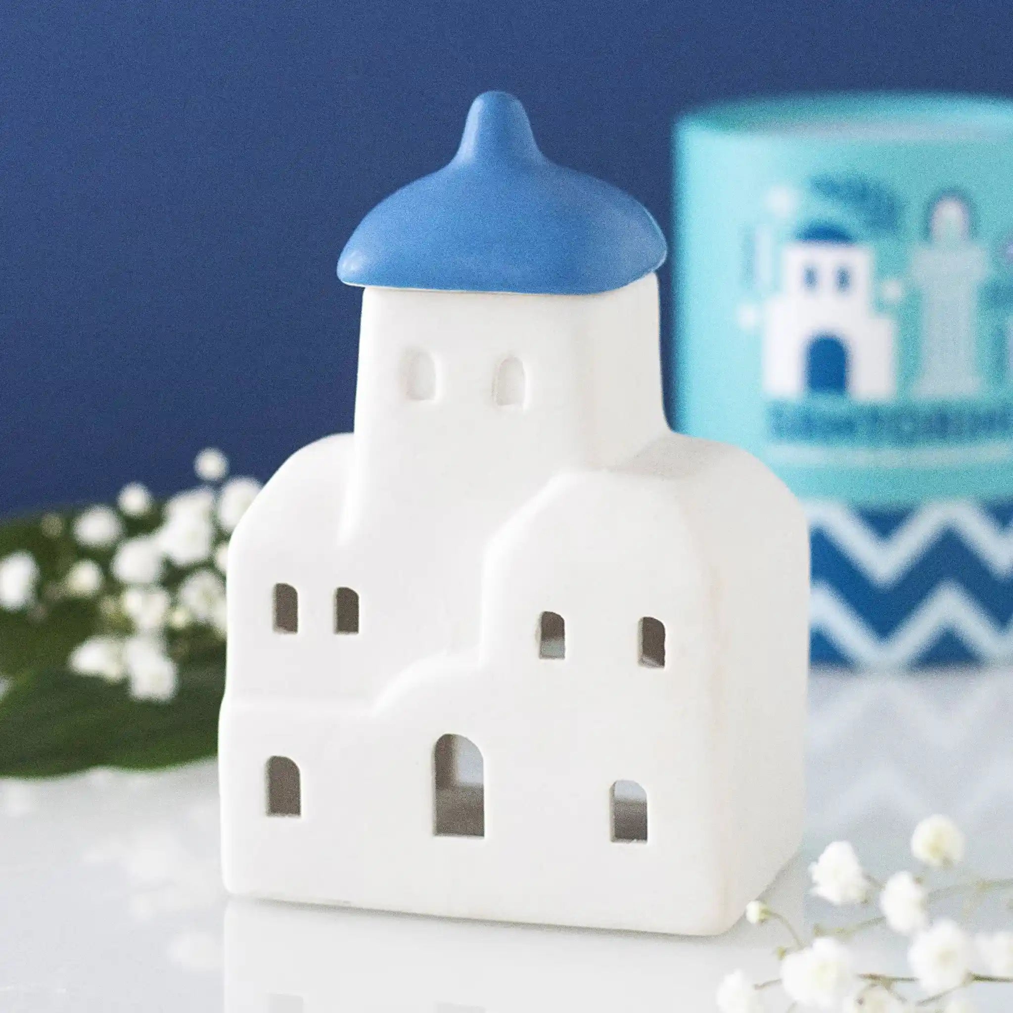 Quemador de aceites esenciales y calentador de cera con forma de casa de Santorini inspirado en Grecia en blanco y azul vista frontal sobre escritorio con decoración al rededor.