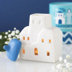 Quemador de aceites esenciales y calentador de cera con forma de casa de Santorini inspirado en Grecia en blanco y azul vista frontal sobre escritorio con decoración al rededor, tapa quitada y vela encendida.