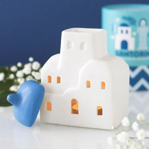 Quemador de aceites esenciales y calentador de cera con forma de casa de Santorini inspirado en Grecia en blanco y azul vista frontal sobre escritorio con decoración al rededor, tapa quitada y vela encendida.