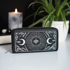 Cartera de cremallera con estampado frontal inspirado en la ouija estilo gótico y witchy vista frontal sobre escritorio con decoración al rededor.