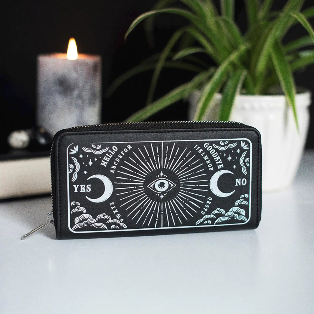 Cartera de cremallera con estampado frontal inspirado en la ouija estilo gótico y witchy vista frontal sobre escritorio con decoración al rededor.