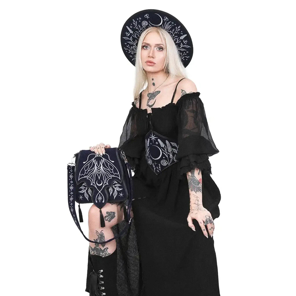 Bolso rígido en azul medianoche con ribetes en negro, bordado en tonos plateados con un diseño celestial de polilla, luna, estrellas y vegetación estilo whimsigoth. Borlas decorativas en la solapa. Vista con modelo sentada