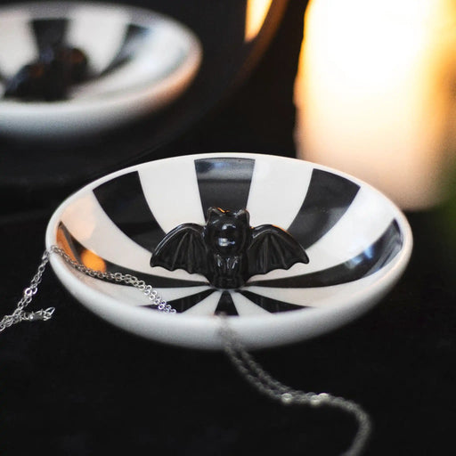 Plato para joyería con rayas negras y murciélago en 3d en el centro visto sobre una mesa con joyería al rededor.