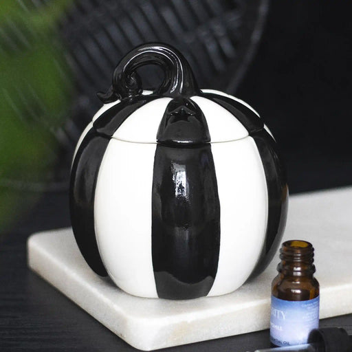 Quemador de aceites esenciales con forma de calabaza a rayas blancas y negras estilo gótico, witchy o alternativo vista frontal sobre escritorio con decoración al rededor.