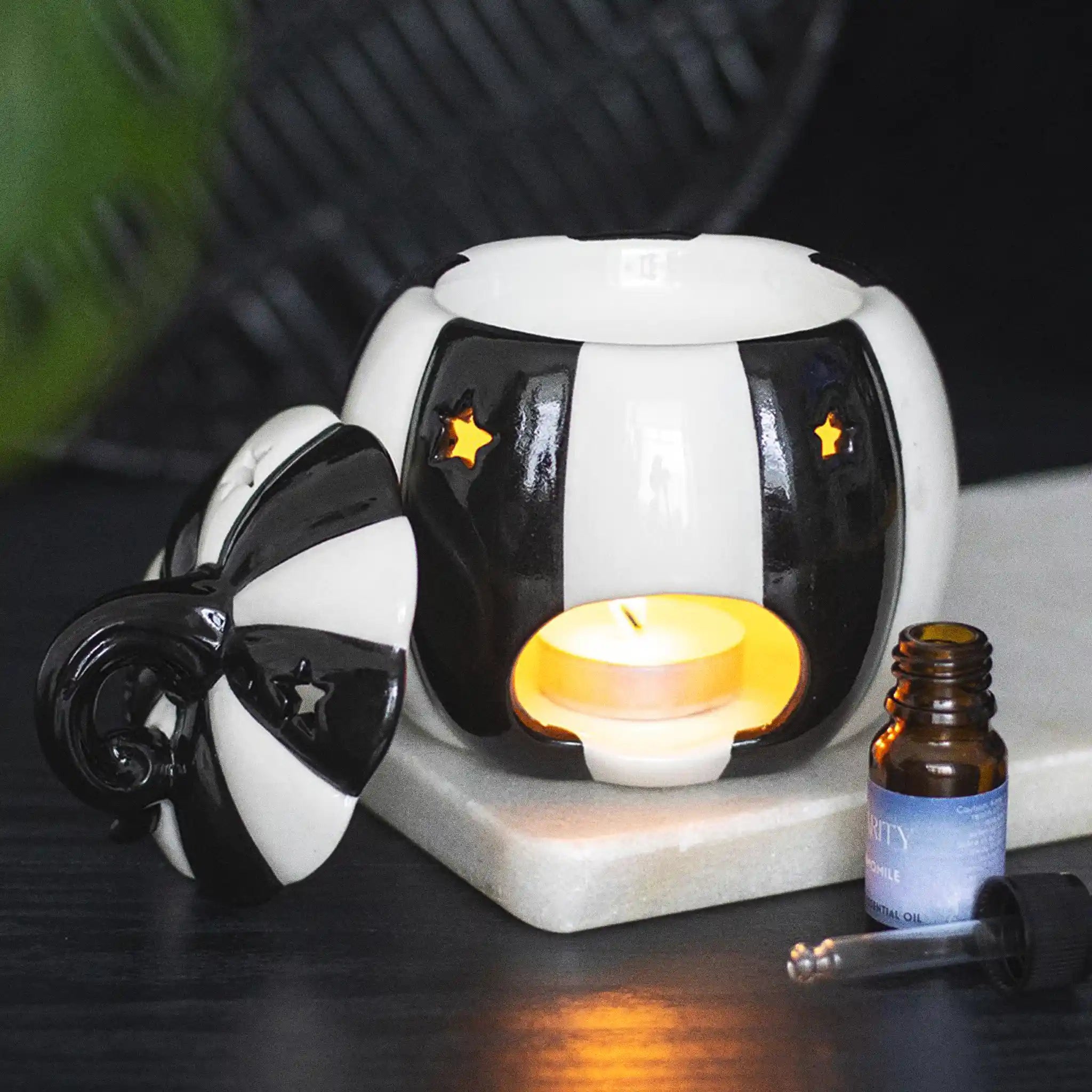 Quemador de aceites esenciales con forma de calabaza a rayas blancas y negras estilo gótico, witchy o alternativo vista posterior con vela encendida sobre escritorio con decoración al rededor.