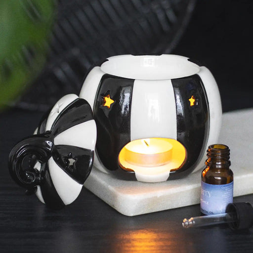 Quemador de aceites esenciales con forma de calabaza a rayas blancas y negras estilo gótico, witchy o alternativo vista posterior con vela encendida sobre escritorio con decoración al rededor.