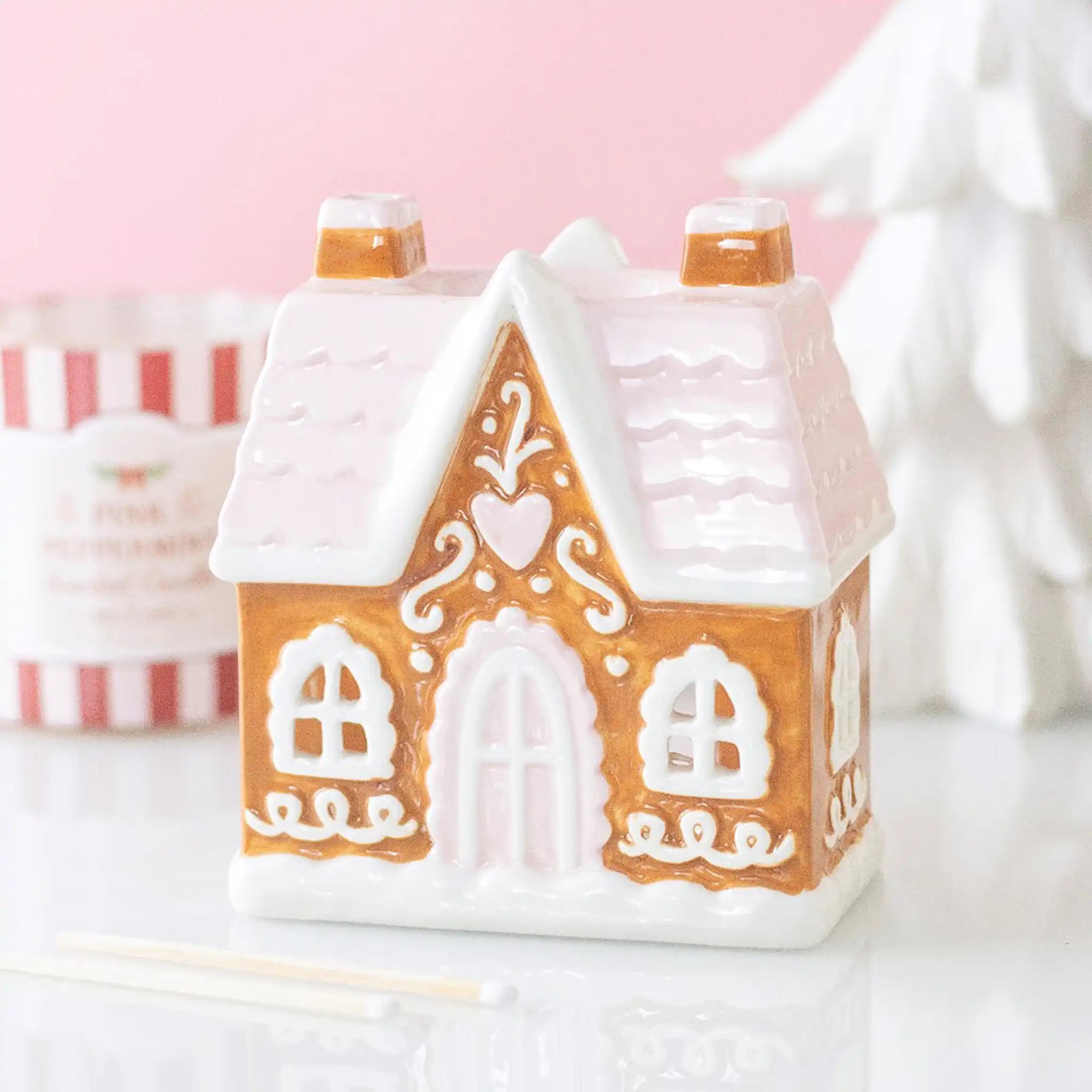 Quemador de aceite y calentador de cera en forma de casita de jengibre estilo navideño en marrón, blanco y rosa vista sobre escritorio con decoración al rededor.