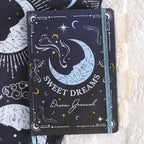 Libreta A5 apoyada sobre una manta con portada de ilustración de lunas y estrellas.