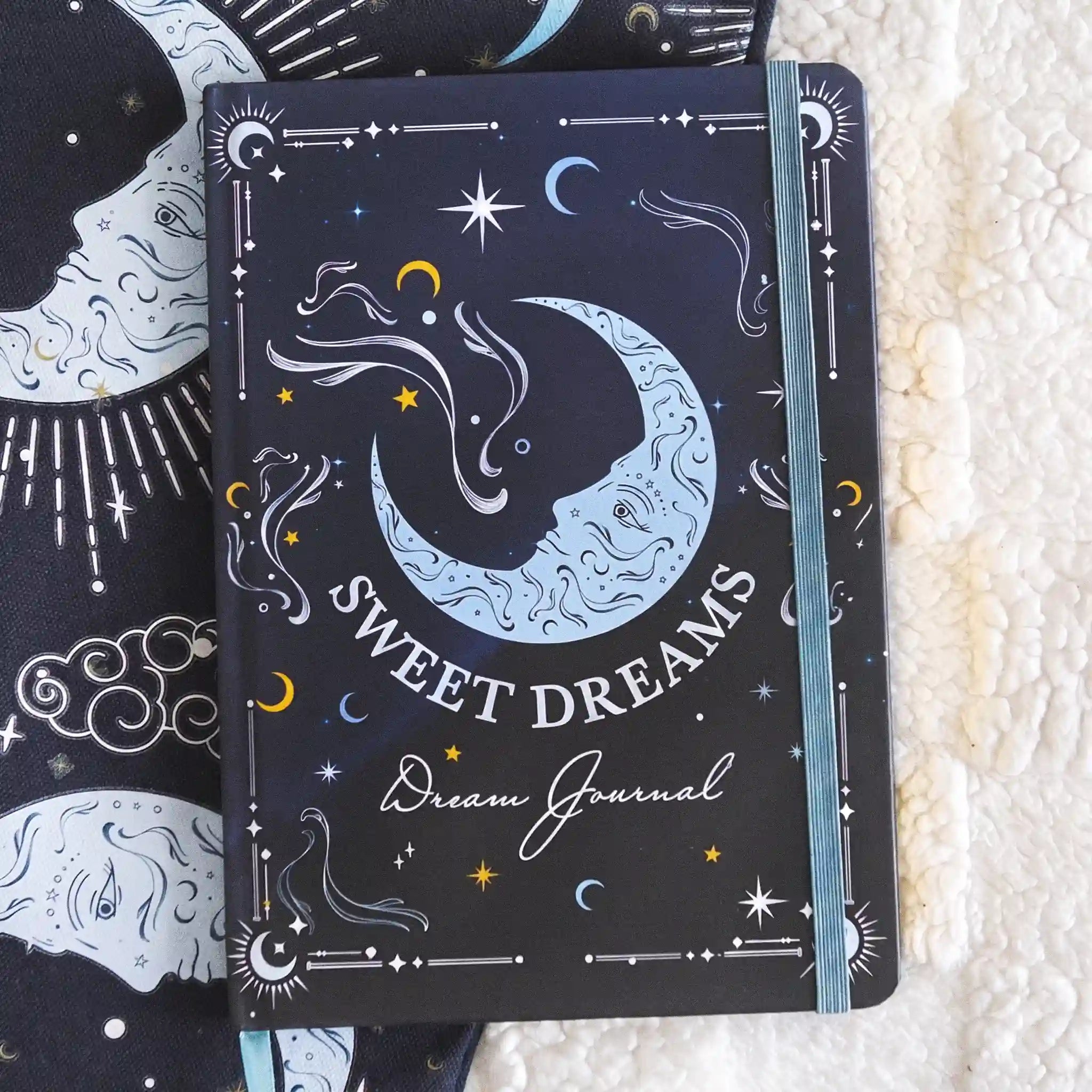 Libreta A5 apoyada sobre una manta con portada de ilustración de lunas y estrellas.