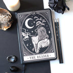 Libreta para lectoras de fantasía y romantasy con dragón, luna y chica leyendo en la portada y cinta marcapáginas en estética de carta de tarot color blanco y negro vista sobre escritorio con decoración al rededor.