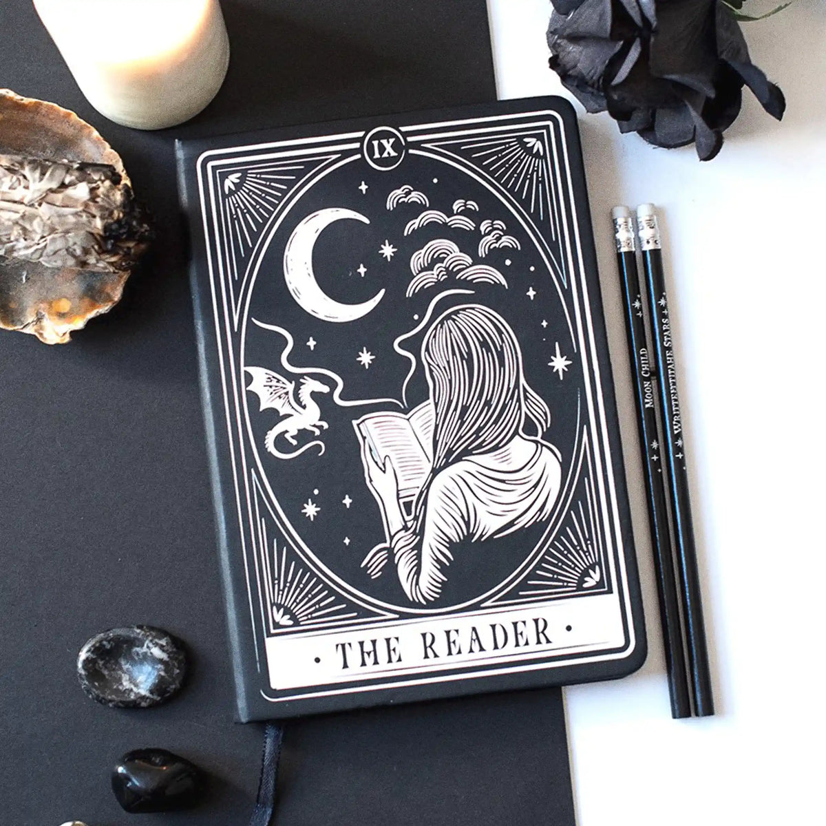 Libreta para lectoras de fantasía y romantasy con dragón, luna y chica leyendo en la portada y cinta marcapáginas en estética de carta de tarot color blanco y negro vista sobre escritorio con decoración al rededor.