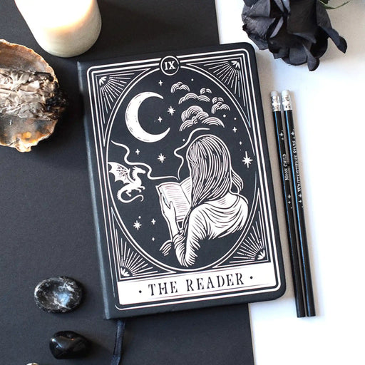 Libreta para lectoras de fantasía y romantasy con dragón, luna y chica leyendo en la portada y cinta marcapáginas en estética de carta de tarot color blanco y negro vista sobre escritorio con decoración al rededor.