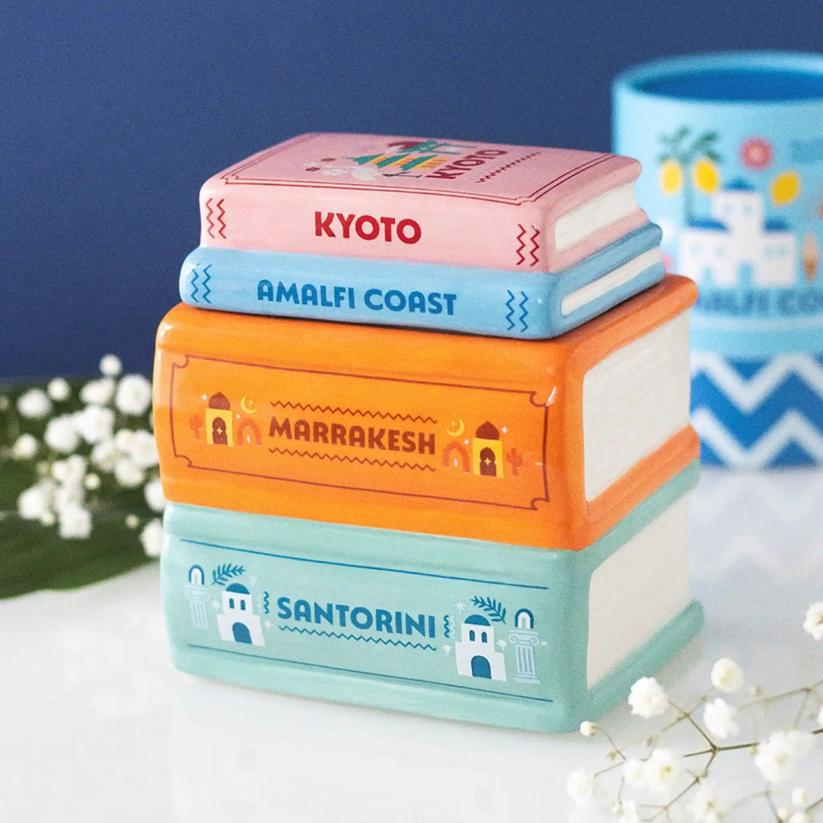 Quemador de aceites esenciales y calentador de cera en forma de libros apilados de diferentes destinos de viaje multicolor vista frontal sobre escritorio con decoración al rededor.