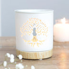 Quemador de aceites esenciales y calentador de cera con diseño de árbol de la vida calado sobre cerámica y base de madera vista frontal sobre escritorio con decoración al rededor.