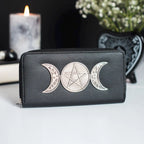 Cartera con adorno metálico frontal de triple luna y pentagrama sobre piel sintética negra con cierre de cremallera vista sobre escritorio con decoración al rededor.