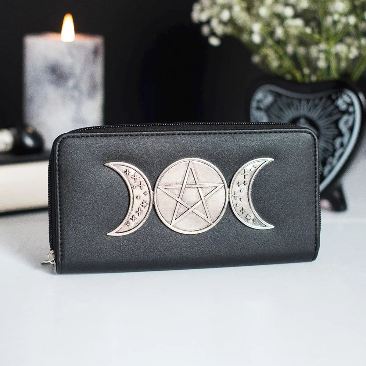 Cartera con adorno metálico frontal de triple luna y pentagrama sobre piel sintética negra con cierre de cremallera vista sobre escritorio con decoración al rededor.