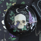 Plato para joyería con diseño de calavera, serpiente y plantas estilo gótico sobre mesa con joyería al rededor.