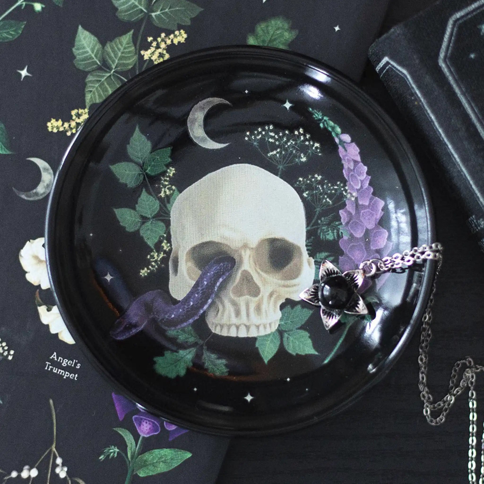 Plato para joyería con diseño de calavera, serpiente y plantas estilo gótico sobre mesa con joyería al rededor.