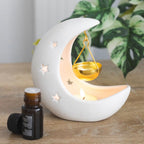 Quemador de aceites esenciales en forma de luna creciente en color blanco con estrellas cut out y recipiente colgante para aceites en color dorado vista sobre escritorio con decoración al rededor. 