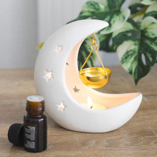 Quemador de aceites esenciales en forma de luna creciente en color blanco con estrellas cut out y recipiente colgante para aceites en color dorado vista sobre escritorio con decoración al rededor. 