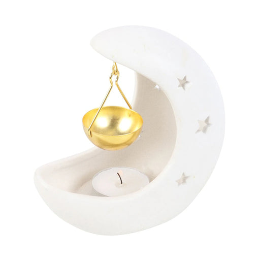 Quemador de aceites esenciales en forma de luna creciente en color blanco con estrellas cut out y recipiente colgante para aceites en color dorado vista con vela sobre fondo blanco.