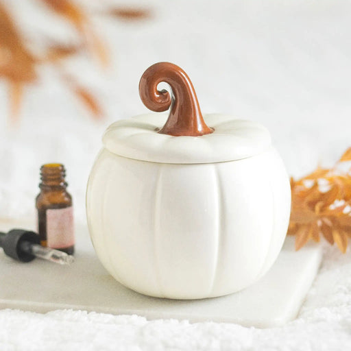Quemador de aceites esenciales de cerámica con forma de calabaza con tallo rizado en blanco roto y marrón estilo aesthetic para otoño y halloween vista frontal sobre escritorio con decoración al rededor.