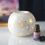Quemador de aceites esenciales y calentador de ceras aromáticas con forma de bola con estrellas caladas en color blanco iridiscente vista frontal sobre escritorio con decoración al rededor.