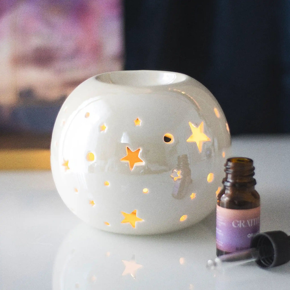 Quemador de aceites esenciales y calentador de ceras aromáticas con forma de bola con estrellas caladas en color blanco iridiscente vista frontal sobre escritorio con decoración al rededor.