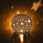 Quemador de aceites esenciales y calentador de ceras aromáticas con forma de bola con estrellas caladas en color blanco iridiscente vista frontal con vela encendida dentro.