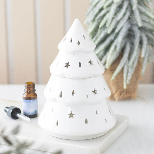 Quemador de aceites esenciales con forma de árbol de navidad y estrellas y gotas cut out en la superficie vista frontal sobre escritorio con decoración al rededor.