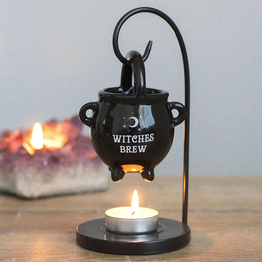 Quemador de aceites esenciales y calentador de cera con forma de caldero de bruja colgado sobre un soporte donde va la vela de té vista frontal sobre escritorio con decoración al rededor.